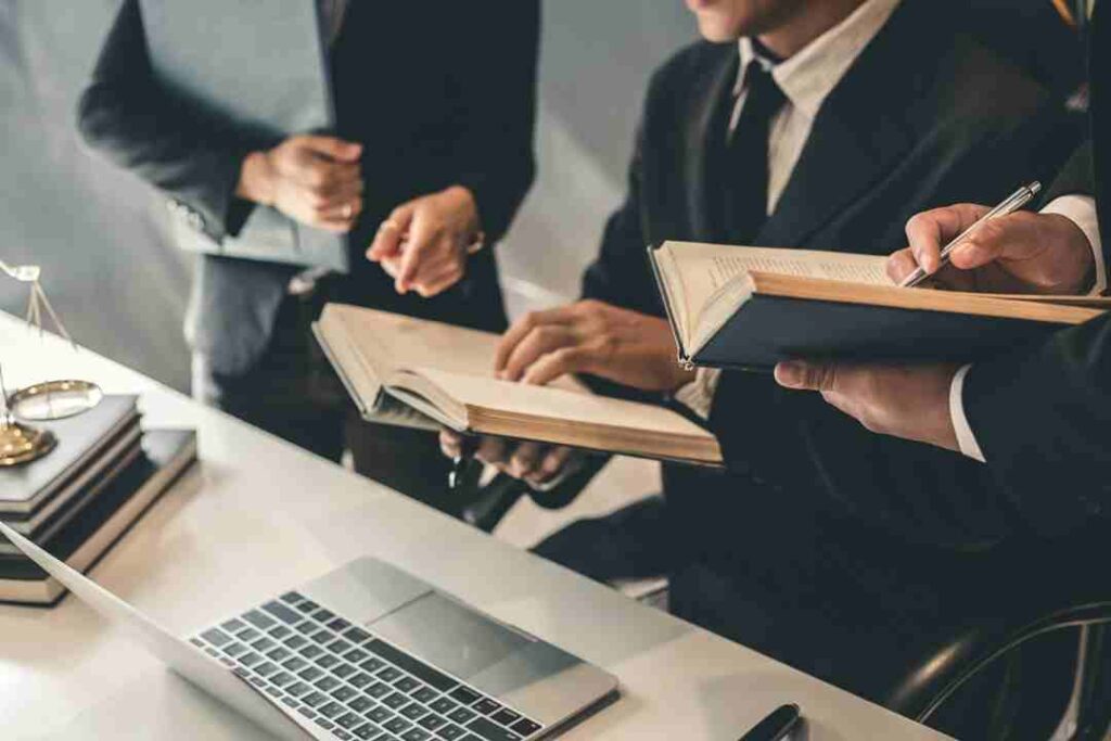 Lawyers in suits discussing books and looking at a laptop.